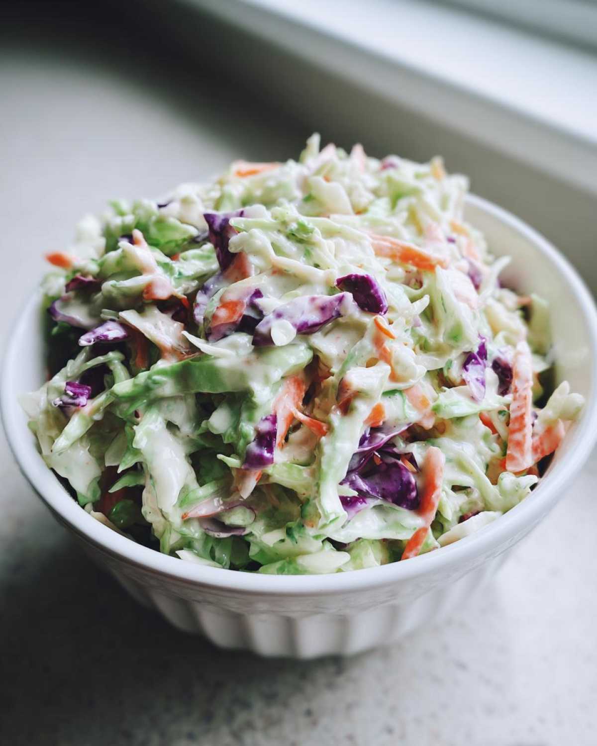 A close-up of a white bowl filled with vibrant Southern Style Creamy Coleslaw, featuring shredded green and purple cabbage and carrots in a rich dressing.