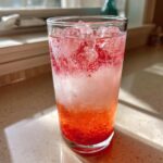 Close-up of a tall glass filled with Sparkling Fresh Strawberry Lemonade, ice, and muddled fruit.