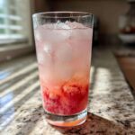 A tall glass filled with Sparkling Fresh Strawberry Lemonade, ice cubes, and muddled strawberries at the bottom, sitting on a granite countertop.