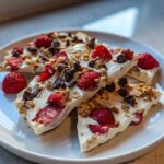 Slices of white Frozen Yogurt Bark topped with fresh strawberries, chocolate chips, and granola on a white plate.