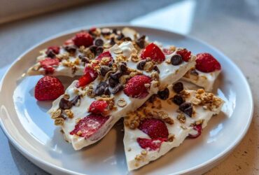 Slices of white Frozen Yogurt Bark topped with fresh strawberries, chocolate chips, and granola on a white plate.