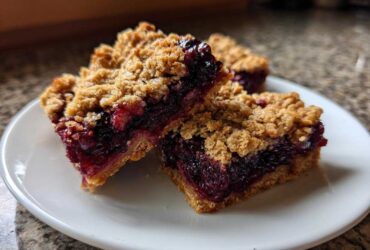 Three delicious Sweet Blackberry Crumble Bars stacked slightly on a white plate, showing the thick blackberry filling and oat topping.
