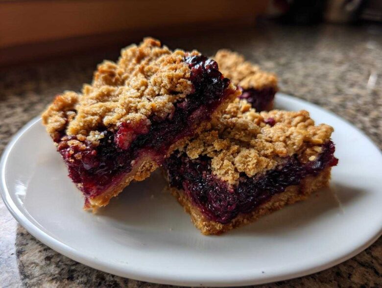 Three delicious Sweet Blackberry Crumble Bars stacked slightly on a white plate, showing the thick blackberry filling and oat topping.
