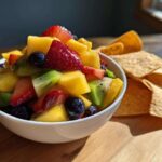 A white bowl filled with Sweet Fresh Fruit Salsa next to a pile of cinnamon tortilla chips on a wooden board.