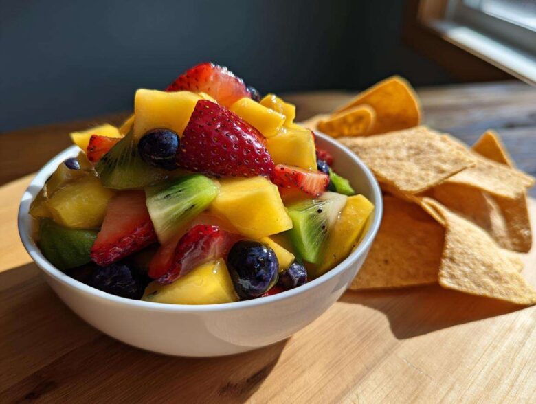 A white bowl filled with Sweet Fresh Fruit Salsa next to a pile of cinnamon tortilla chips on a wooden board.