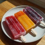 Three vibrant Homemade Fruit Popsicles (red, orange, and purple) resting on a light gray plate.