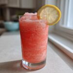 A tall glass filled with bright pink, icy Trendy Frosé, garnished with a lemon wheel.