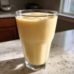 A tall glass filled with a thick, creamy Tropical Pineapple Coconut Smoothie sitting on a light countertop.