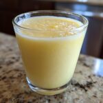Close-up of a bright yellow Tropical Pineapple Coconut Smoothie served in a clear glass on a granite countertop.