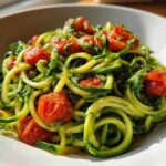 Close-up of a white bowl filled with bright green Zucchini Noodles (Zoodles) tossed in pesto and topped with roasted cherry tomatoes.